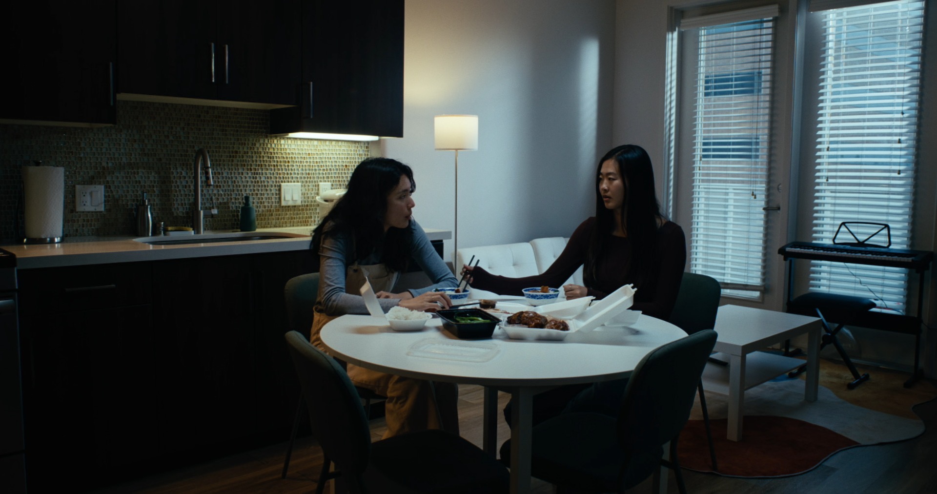 Two women at a round dining table with takeout containers, one passing food across.