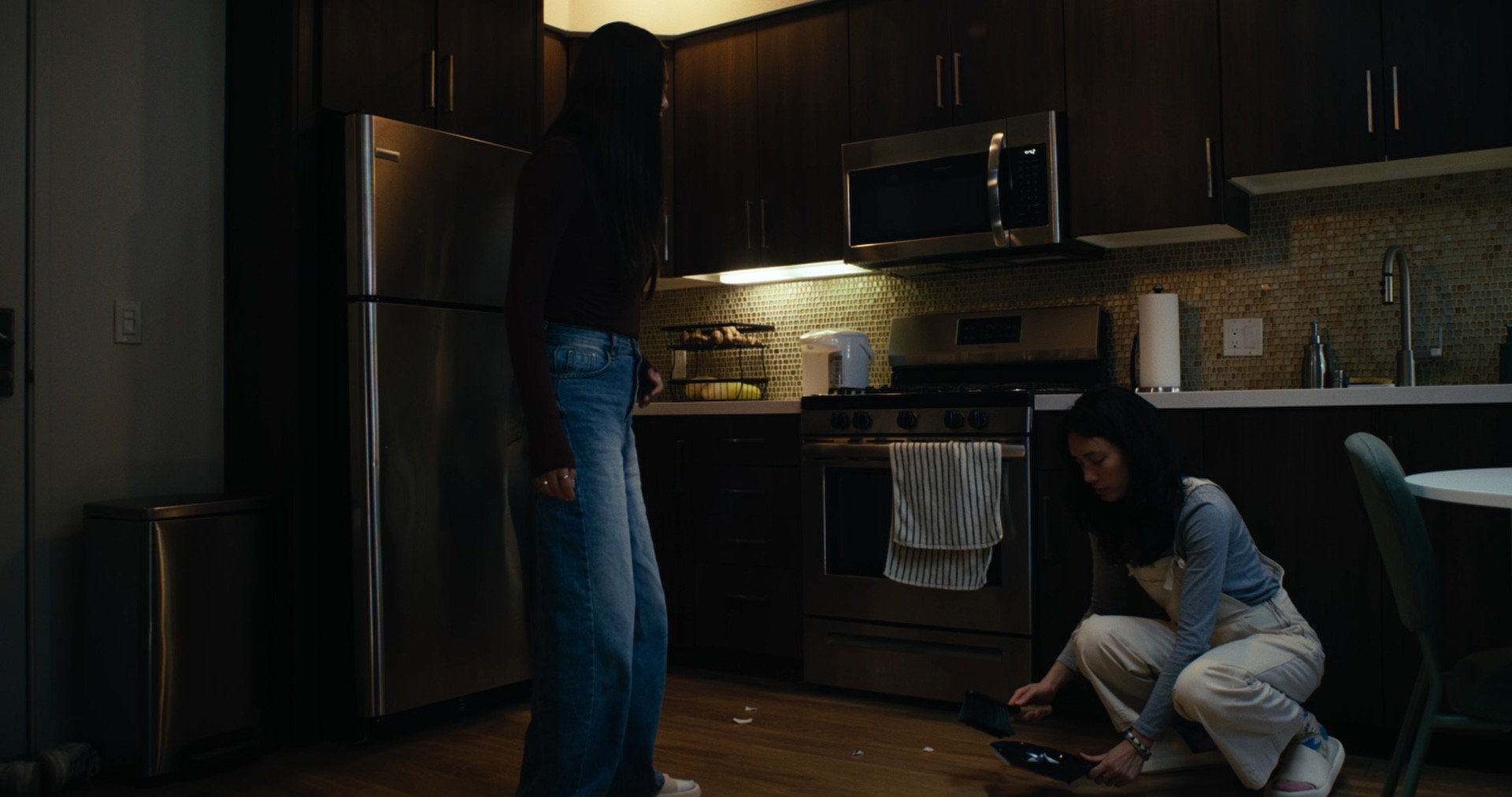 A kitchen: one woman stands while the other crouches to sweep up something broken.